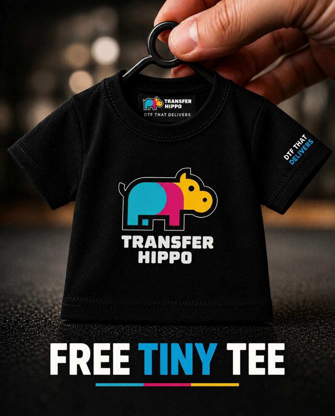 Miniature black Transfer Hippo Tiny Tee with the colorful logo on the front and a printed DTF transfer care-label neck tag reading Transfer Hippo / Size - TINY, held between two fingers